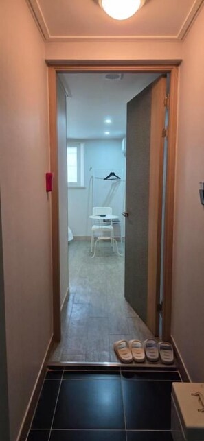 Un pasillo estrecho con una puerta que da a una habitación. Hay zapatillas en el suelo y una caja a la derecha.