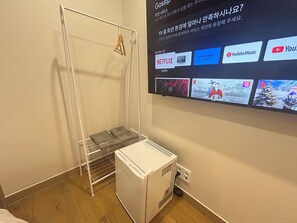 Habitación con televisión de pared blanca que muestra varios servicios de streaming, aire acondicionado blanco y una estantería metálica con ropa doblada.