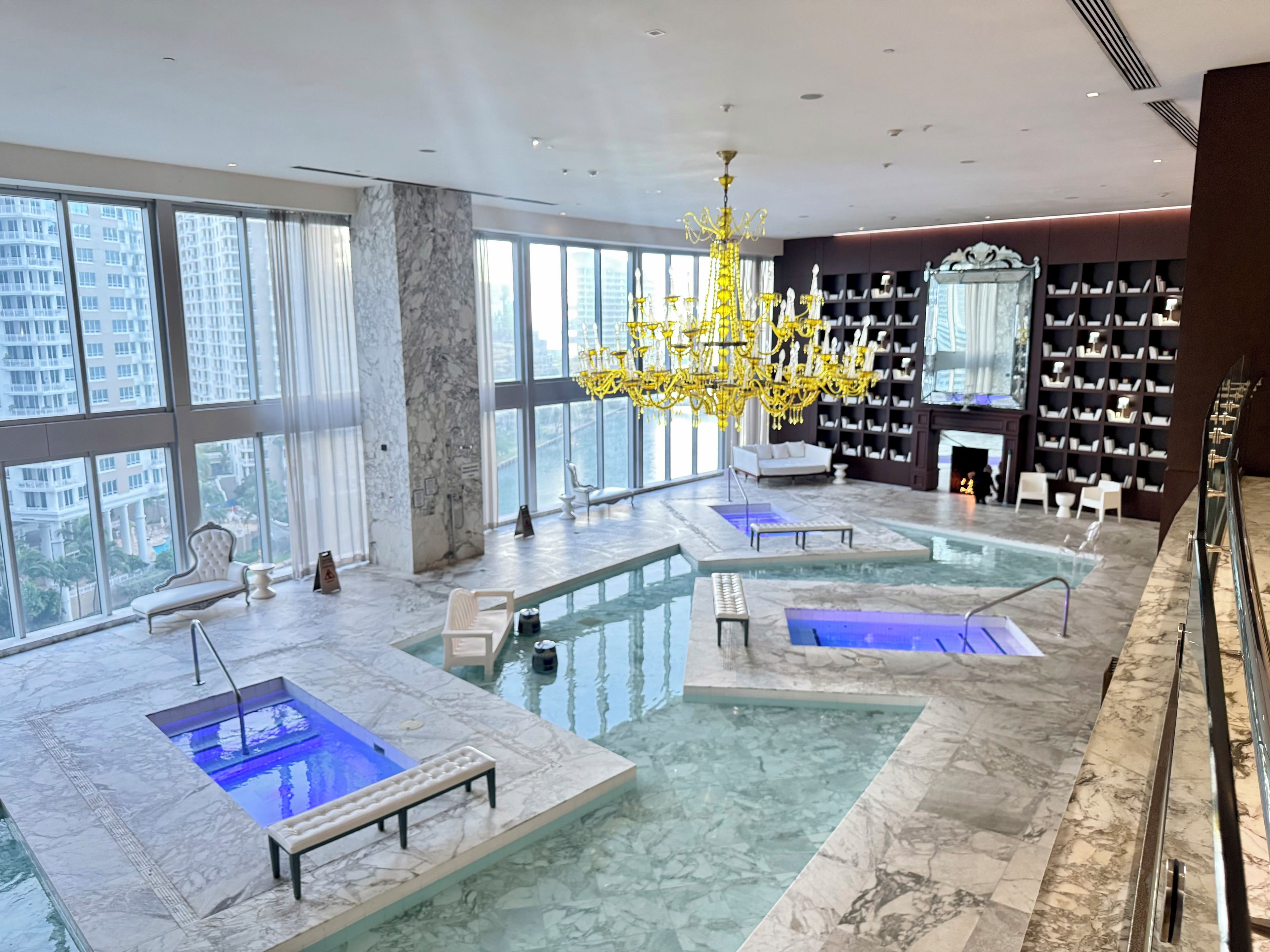 Un'area piscina interna di lusso con diverse vasche idromassaggio, un sontuoso lampadario e vista sulla città attraverso ampie finestre.