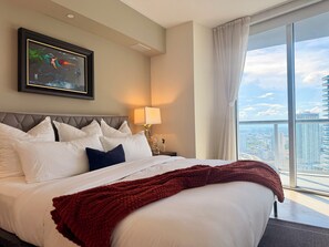 Ein Hotelzimmer mit einem großen Bett, Blick auf die Stadt, einem gerahmten Kunstwerk an der Wand und einem Nachttisch mit Lampe.