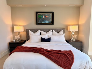 Una cama bien hecha con sábanas blancas y una manta roja, flanqueada por dos mesitas de noche con lámparas y objetos decorativos.