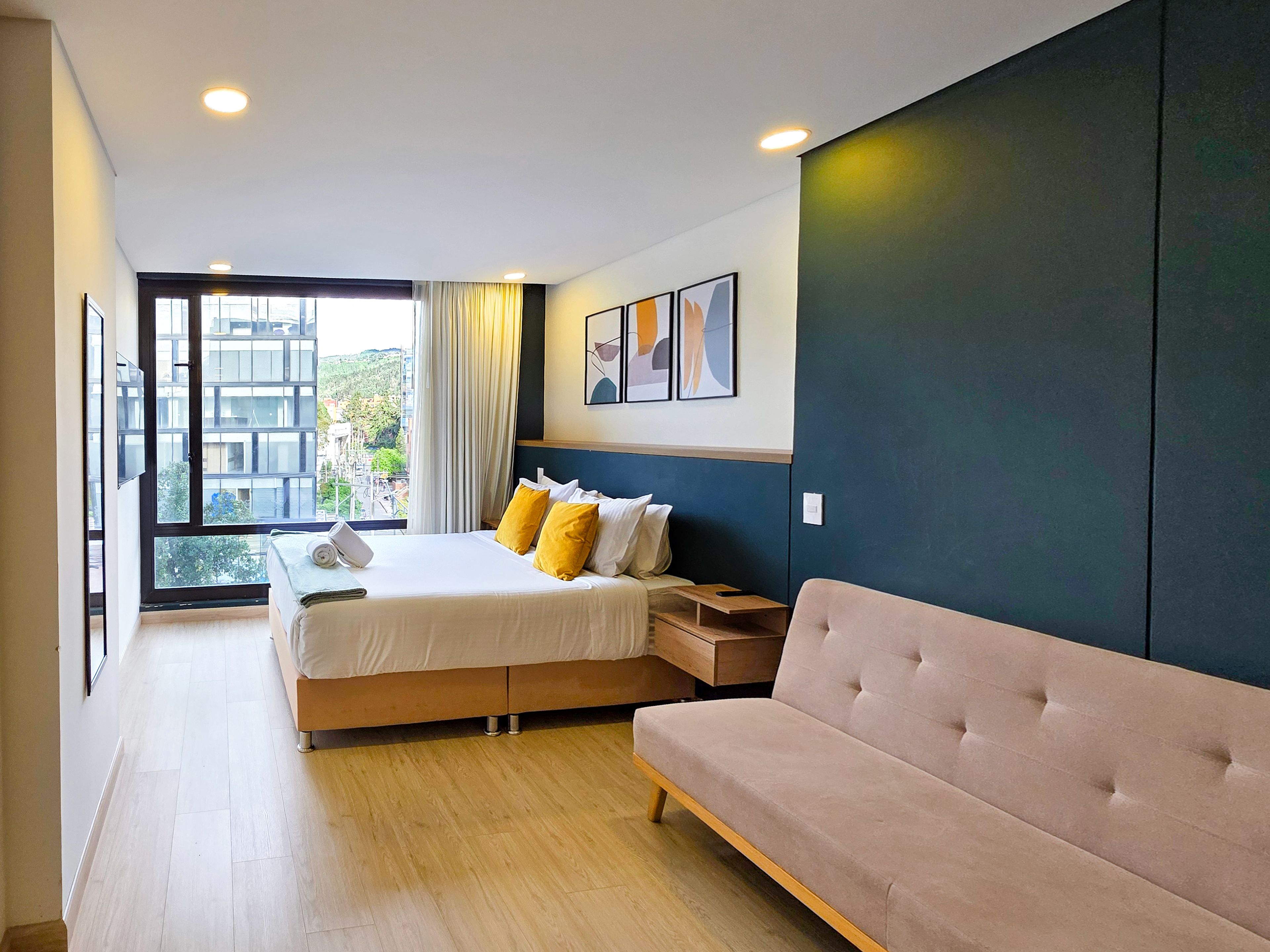 A modern hotel room with a large bed, a sofa, a desk, and a view of the city through a large window.