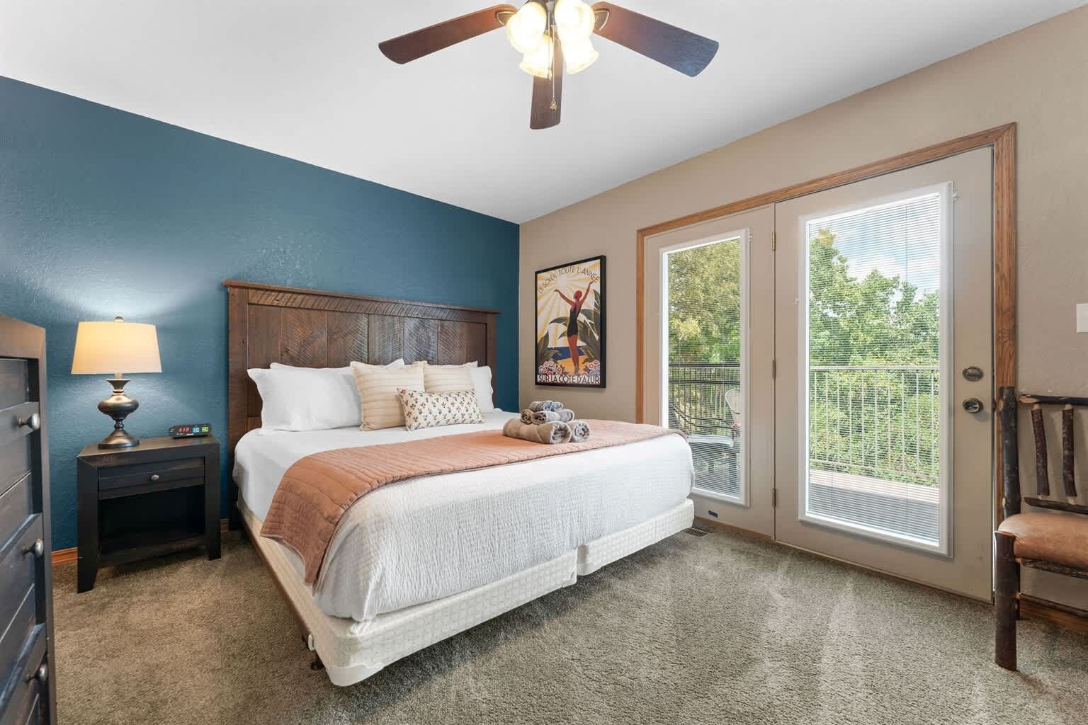A bedroom with a bed, a nightstand with a lamp, a ceiling fan, a window with blinds, and a door leading to a balcony.