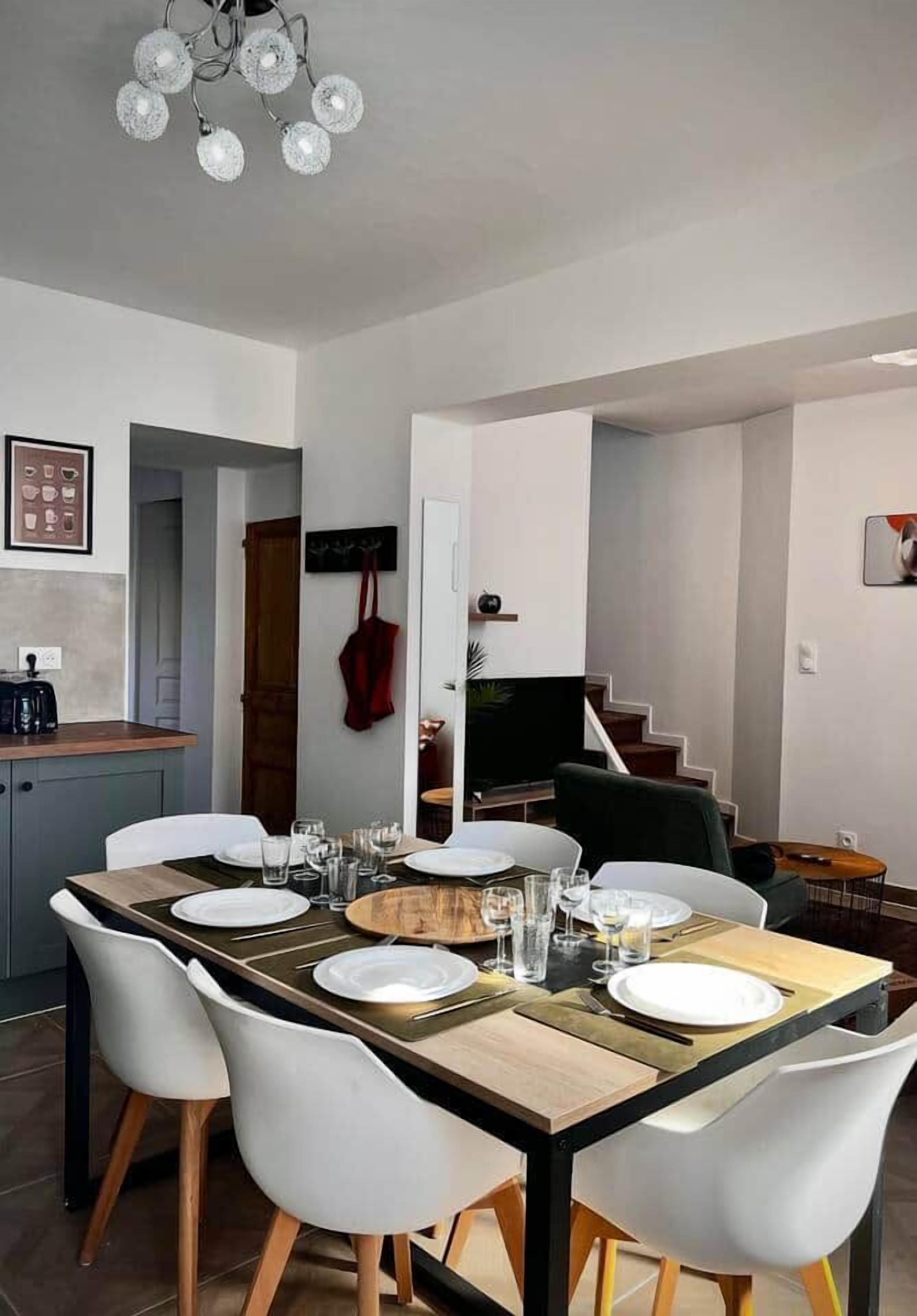 A dining area with a long table set for four, white chairs, and a chandelier above.