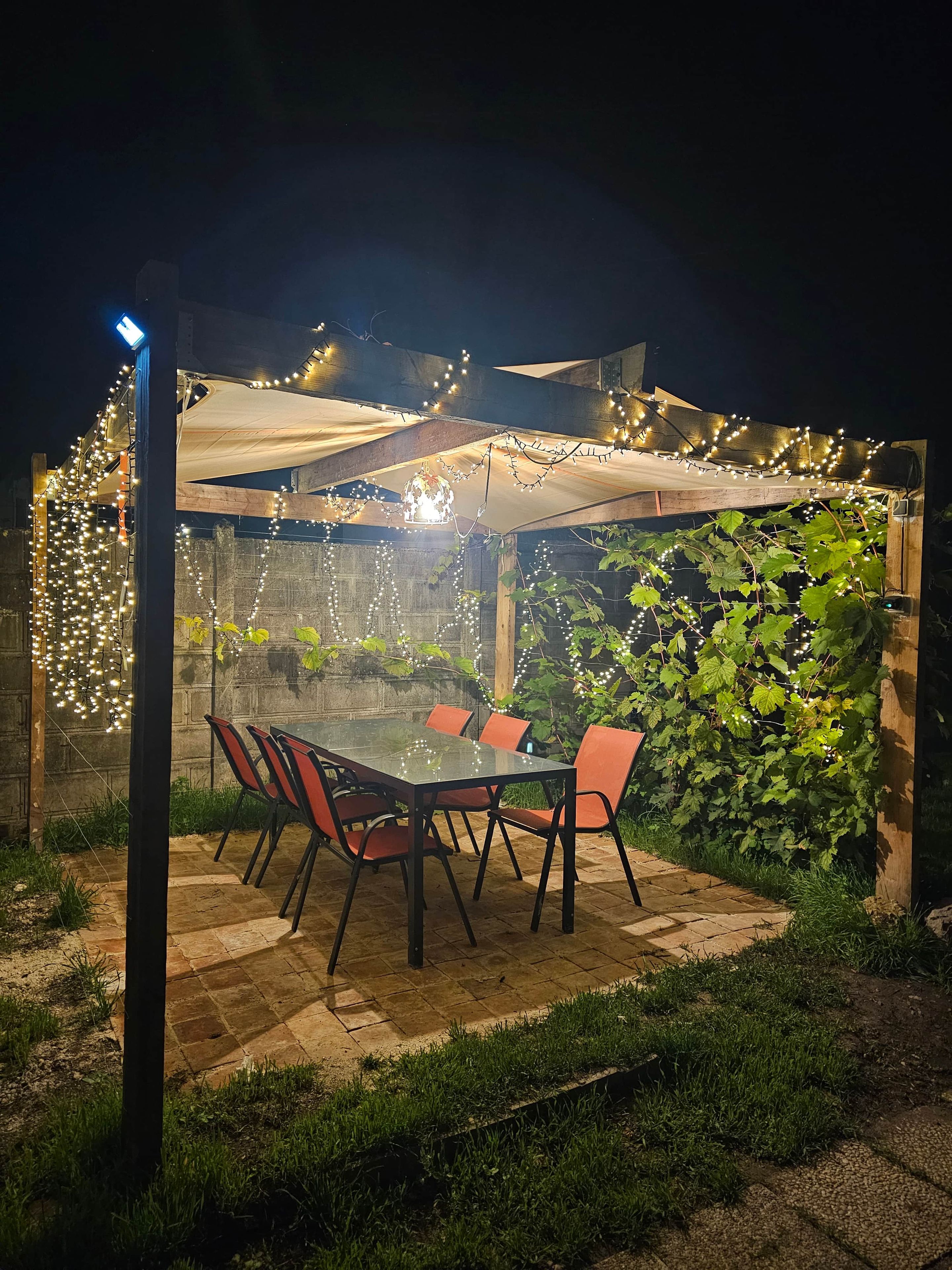 An outdoor patio with a glass table and chairs, surrounded by greenery and string lights.