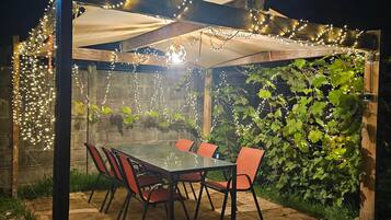 An outdoor patio with a glass table and chairs, surrounded by greenery and string lights.
