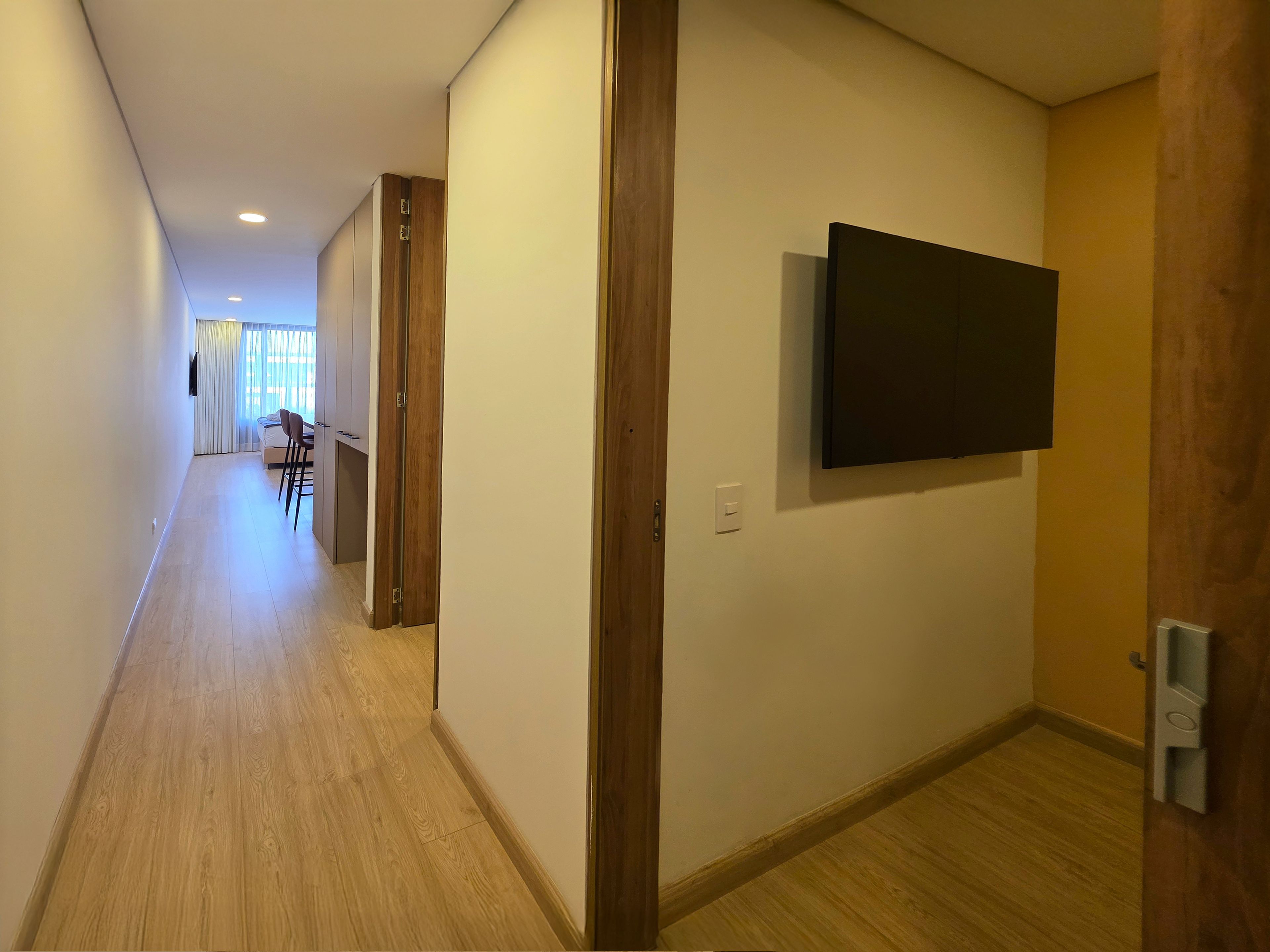 Un pasillo con piso de madera, un televisor de pantalla plana montado en la pared y una puerta que da a otra habitación.