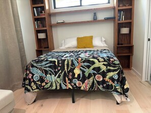 Ein Bett mit einer blumigen Tagesdecke, einem gelben Kopfkissen und Holzregalen mit Büchern und Lampen.