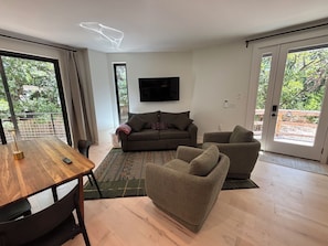 Ein modernes Wohnzimmer mit einem Holzesstisch, einer Couch, einem Sessel und einem an der Wand montierten Fernseher.