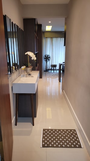 A hallway with a white tiled floor, a small table with a vase of flowers, and a fan in the background. - New Listing (Petrópolis)