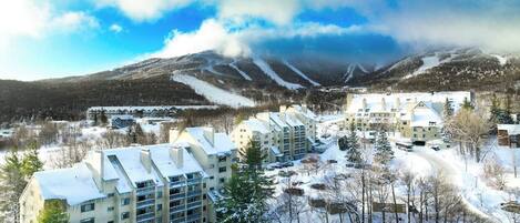 A snow-covered residential area with multi-story buildings, a ski slope in the background, and a clear sky.