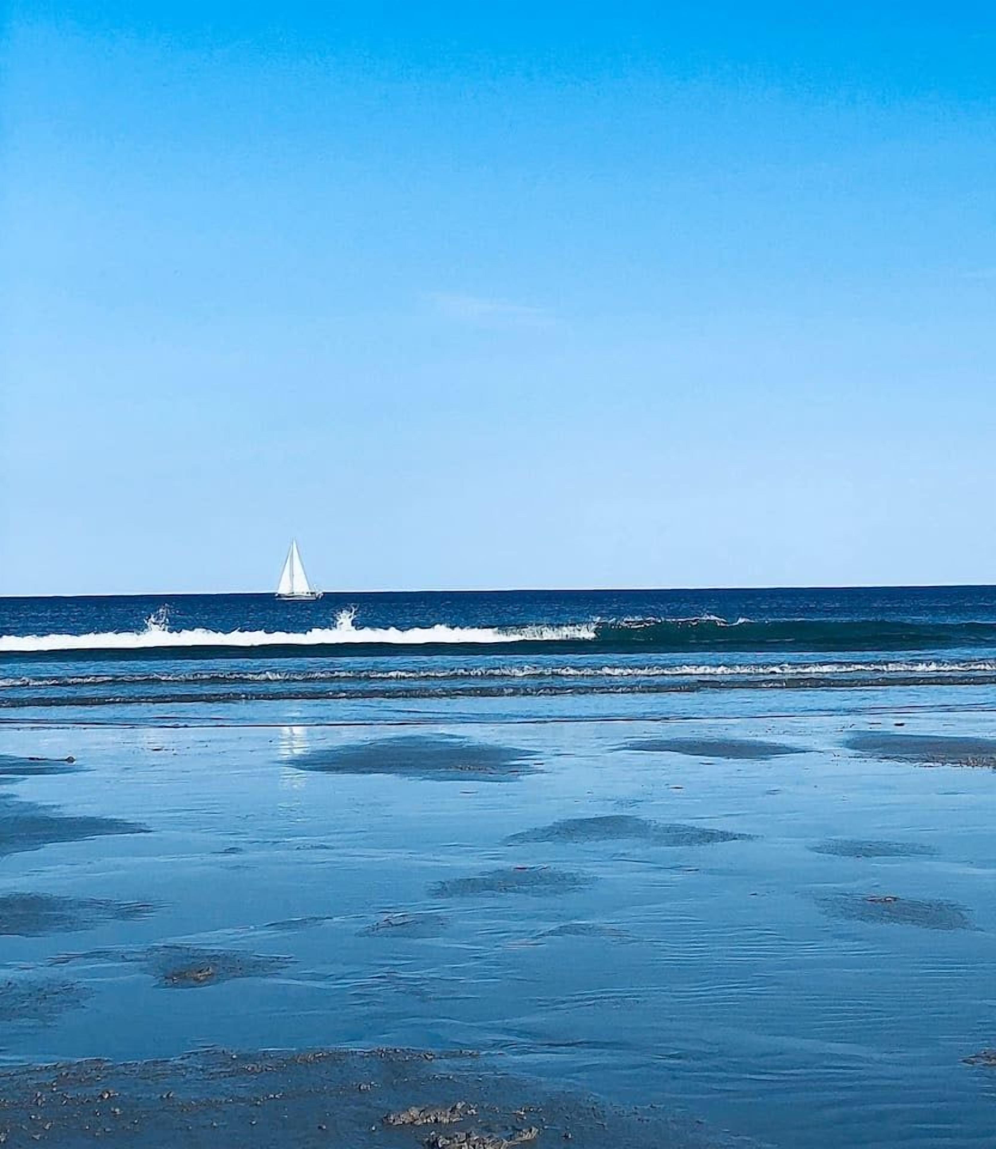 A beach with a sailboat on the water and a clear sky.
