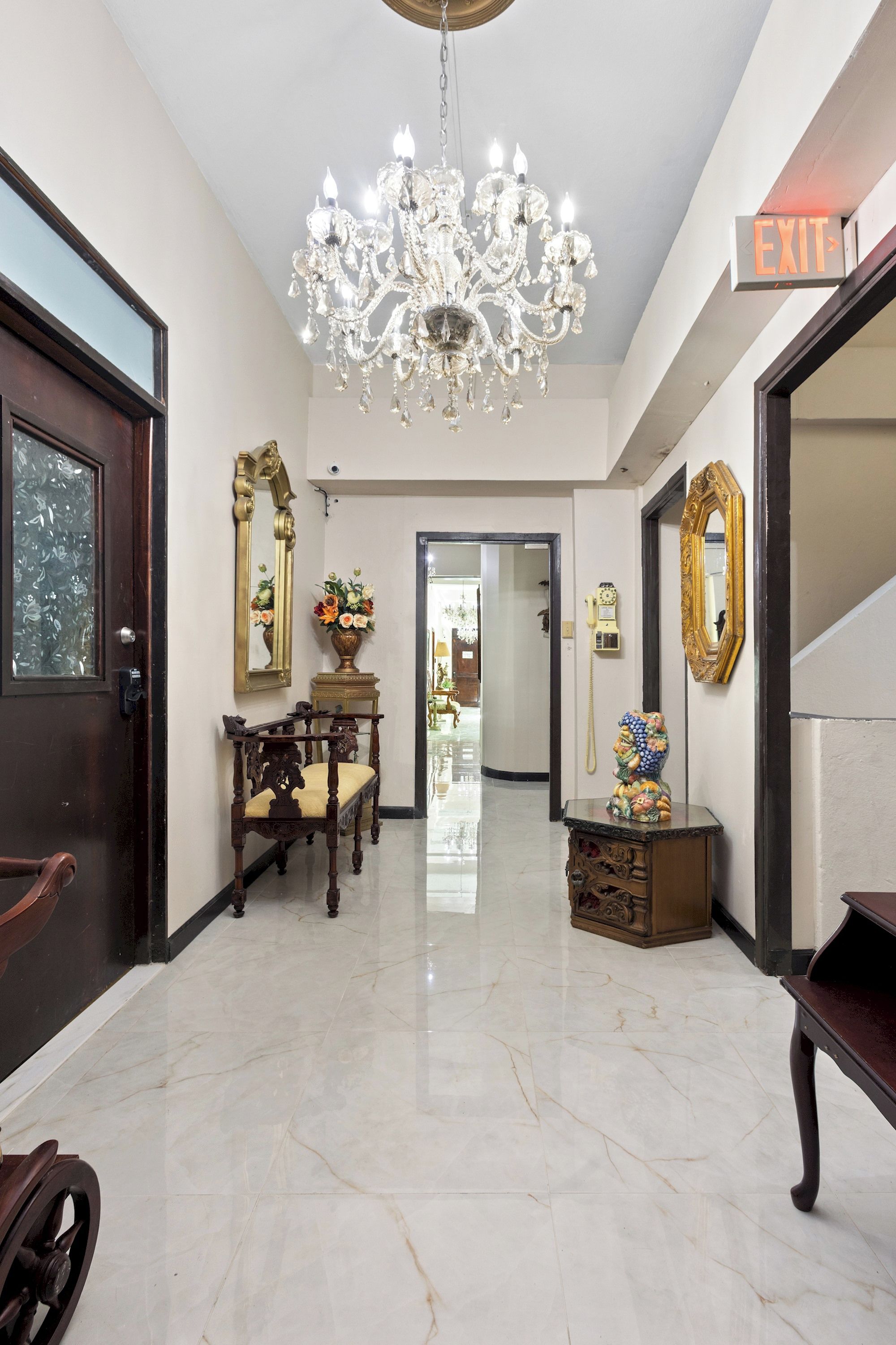 Un pasillo espacioso con piso de mármol, candelabro, banca de madera y una puerta con letrero de 'EXIT'.