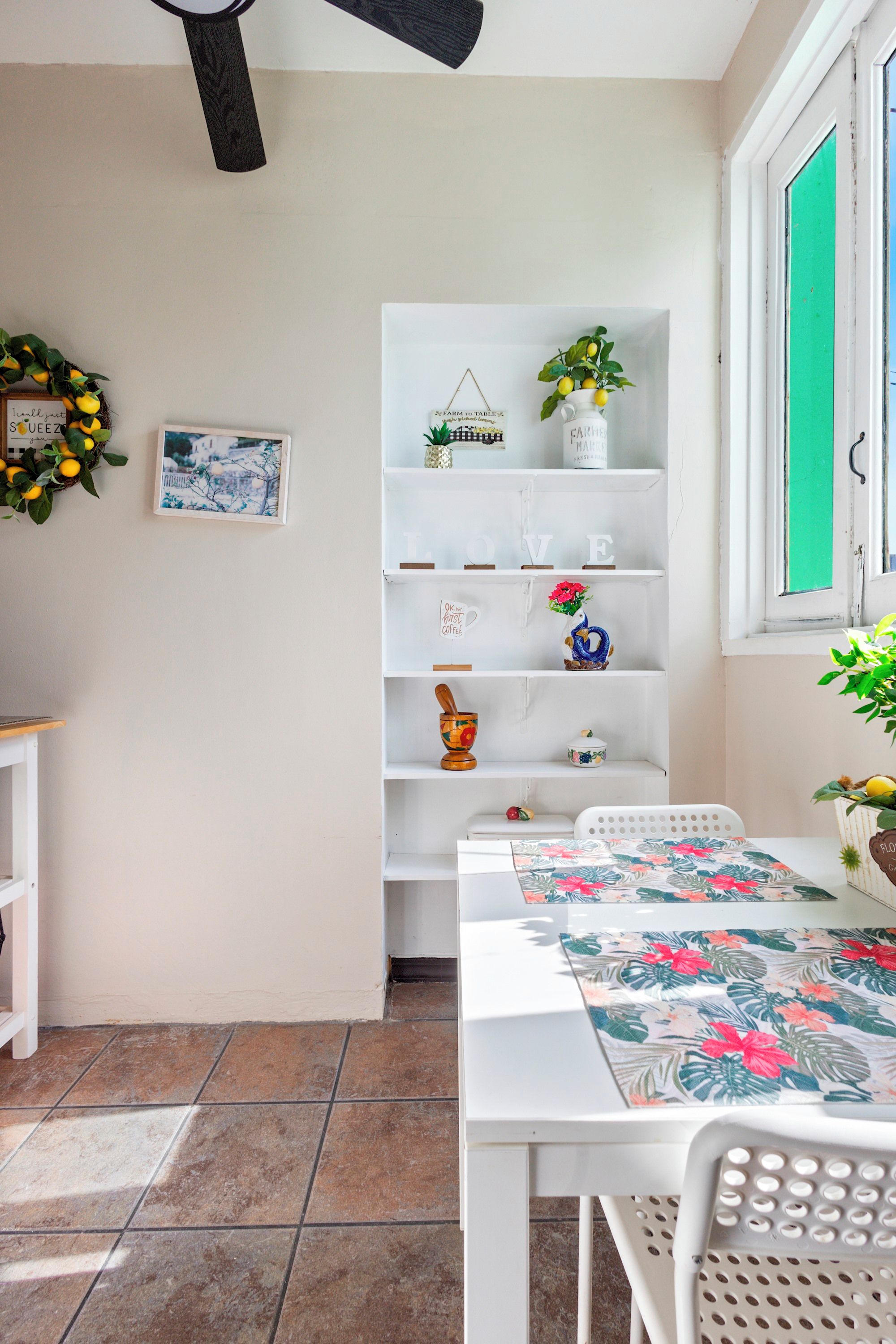Una habitación con una mesa blanca, sillas, un estante con adornos, una ventana que ofrece vistas verdes y un piso con baldosas.