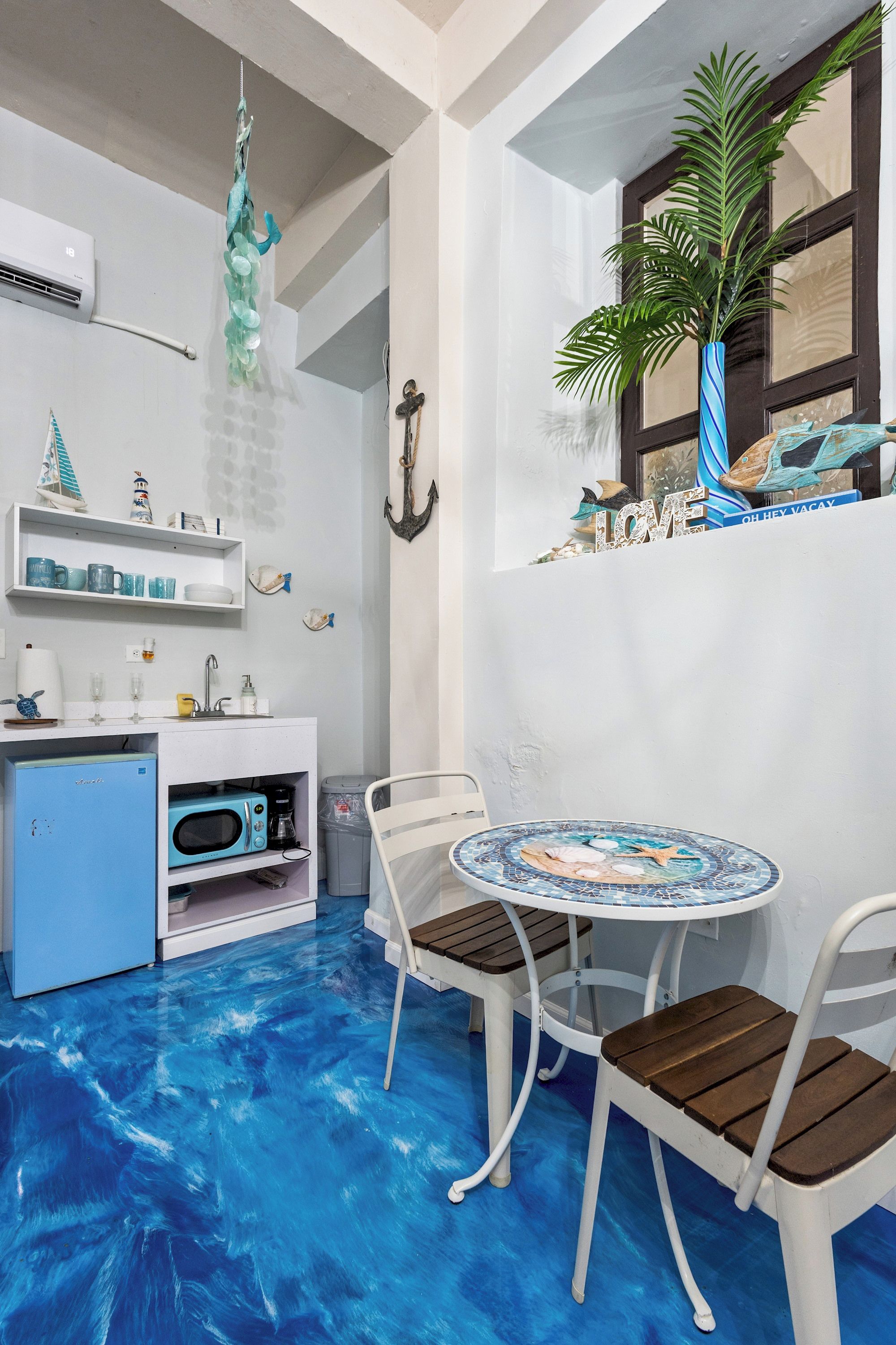 Una habitación con piso azul, paredes blancas, una pequeña zona de cocina, una mesa redonda con sillas y una planta en maceta.