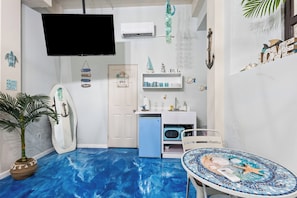 Ein Zimmer mit blauem Boden, einem an der Wand befestigten Fernseher, einem kleinen Tisch mit dekorativer Fliesenplatte und einer Tür mit der Aufschrift 'BEACH'.