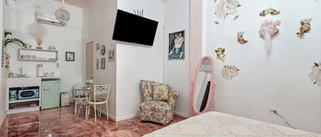Un dormitori amb llit, una cadira, televisió, mirall i una cuineta petita.