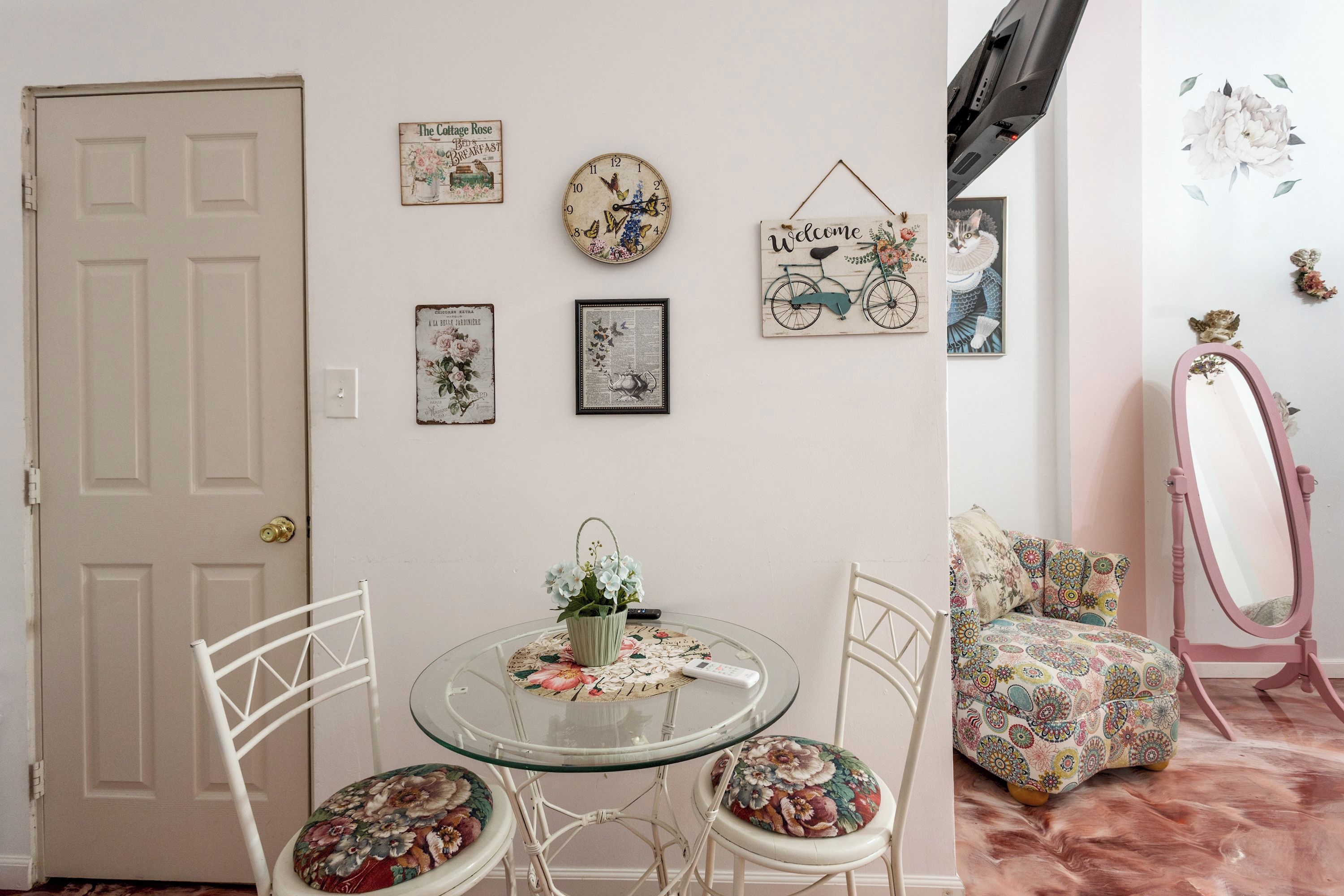 A room with a door, a round glass table with chairs, a wall with various framed pictures, a mirror, and a sofa with patterned cushions.