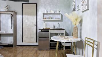 Cocina pequeña con refrigerador, pileta y muebles blancos. Se observa una mesa de comedor con sillas y un jarrón decorativo en la pared.