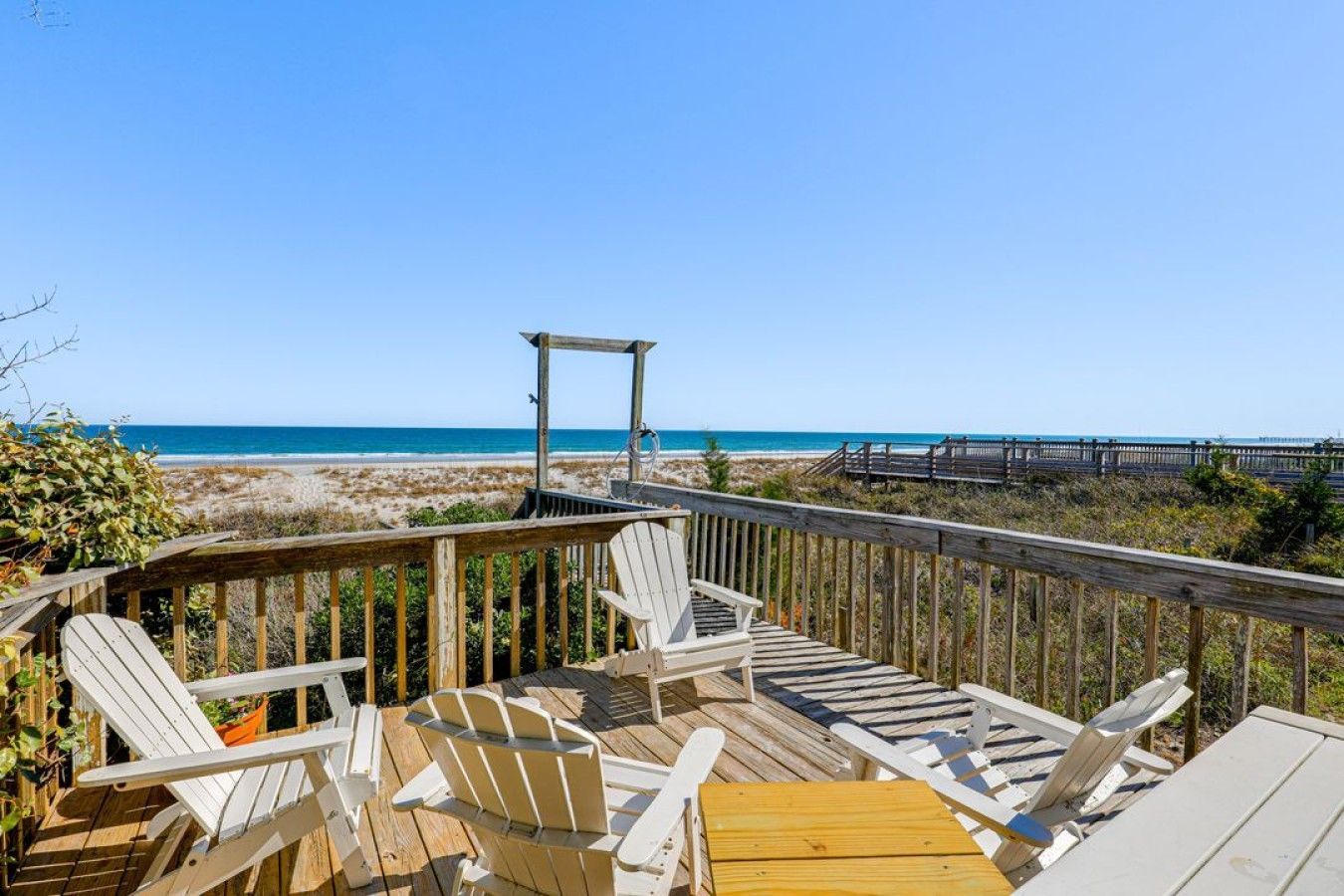 A wooden deck with white Adirondack chairs and a wooden table, overlooking a beach and a boardwalk.