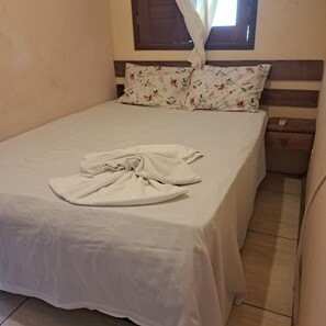 Ein Einzelbett mit weißer Bettdecke und einem gefalteten weißen Bademantel. Am Holzkopfende liegt ein Kissen mit floralem Muster. Ein kleiner Nachttisch mit Fernbedienung ist sichtbar.