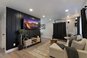 A modern living room with a flat-screen TV mounted on a black wall, a beige sofa, a small round coffee table, and a potted plant. - Private Zion Stay HotTub~Pool~FirePit~Grill+Views (Colorado City)