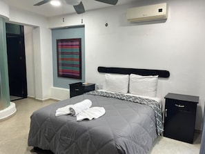 A hotel room with a bed, a nightstand, a small cabinet, a wall-mounted air conditioner, and a colorful tapestry. - New Listing (Palenque)