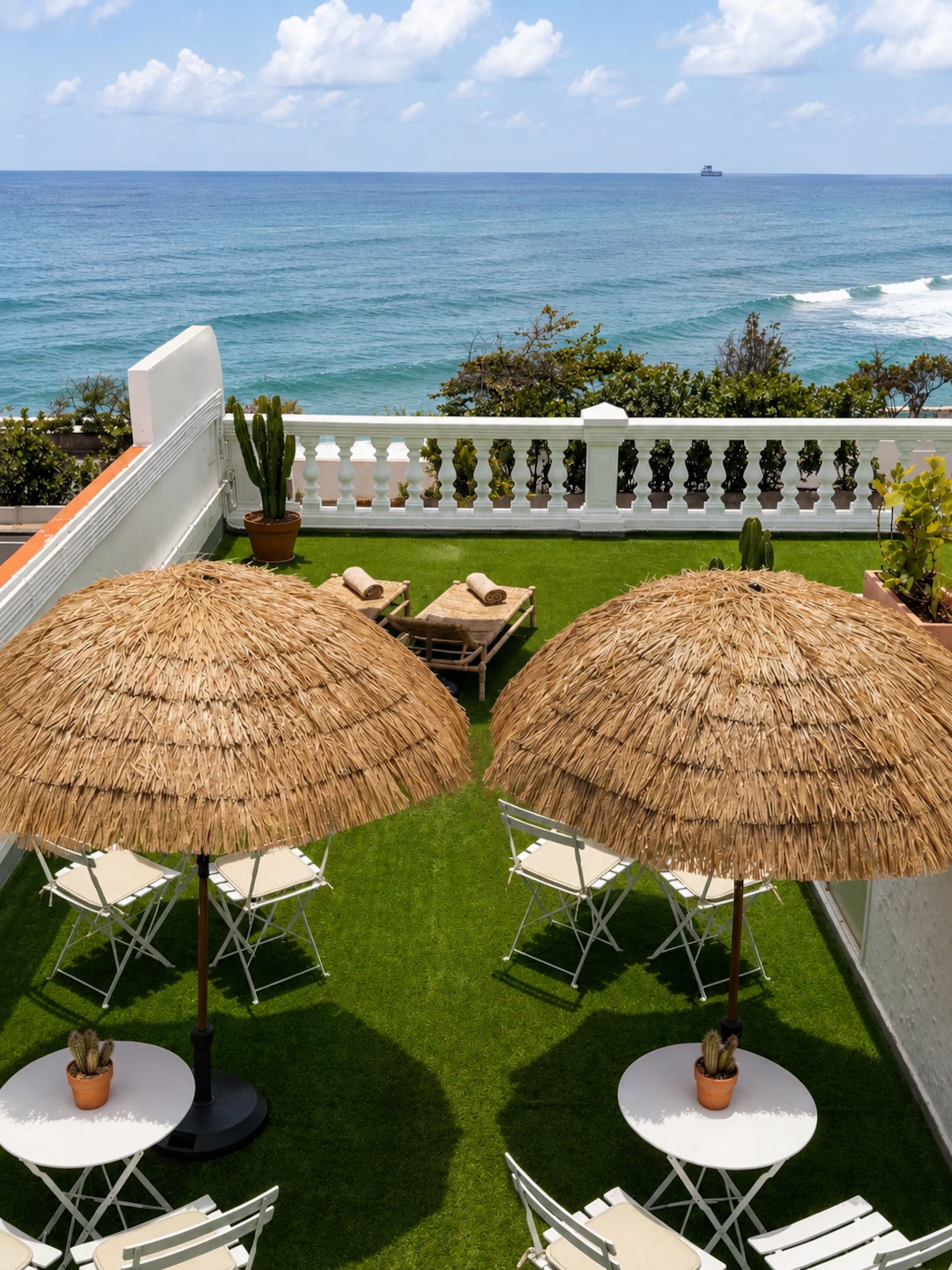 A terrace with two thatched umbrellas, white folding chairs, and a view of the ocean.