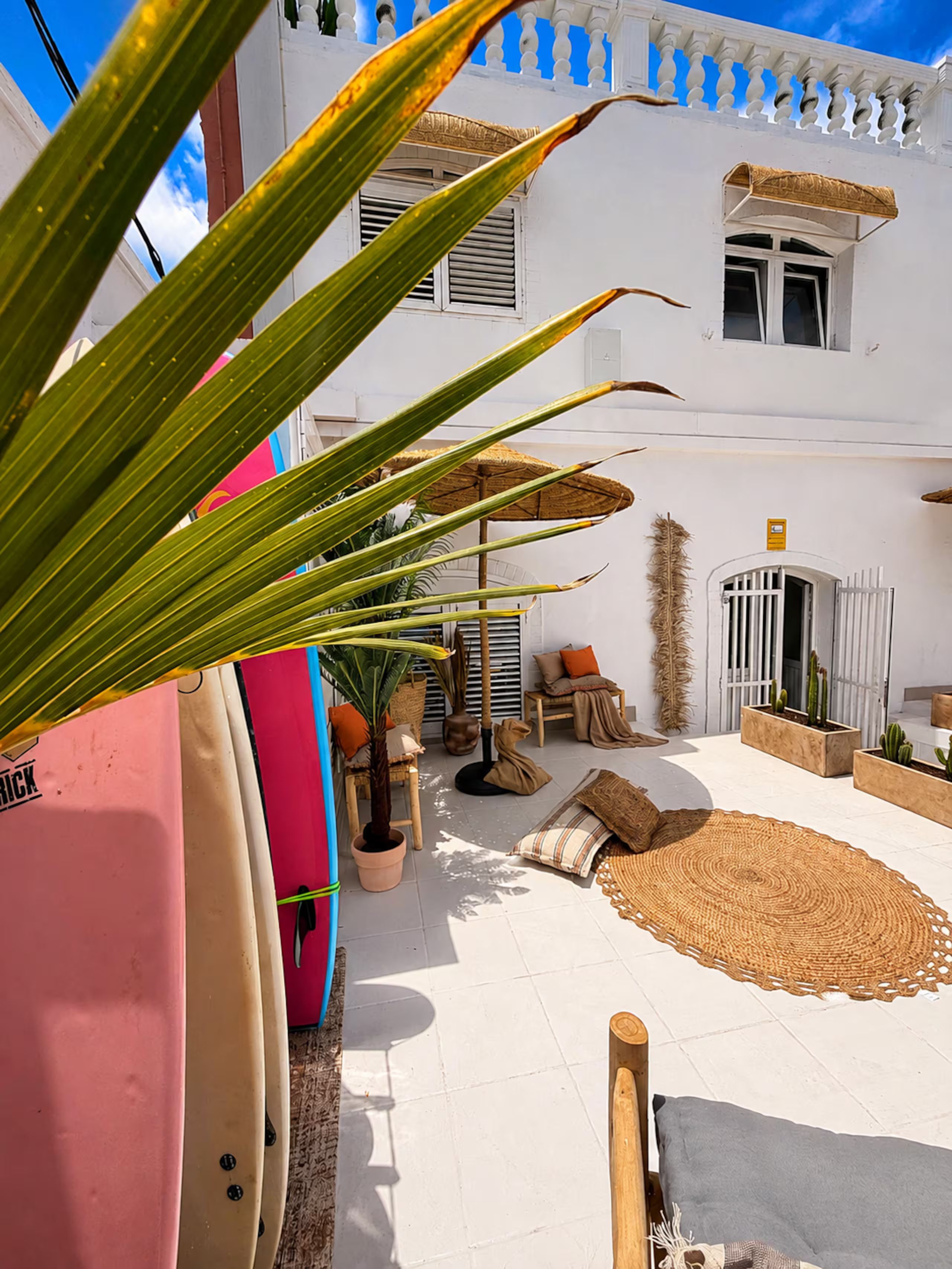 A courtyard with a white building, potted plants, a woven rug, and outdoor furniture.