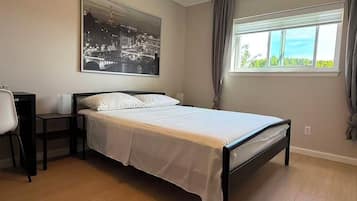 A bedroom with a bed, a desk, a chair, a window with curtains, and a wall-mounted picture.