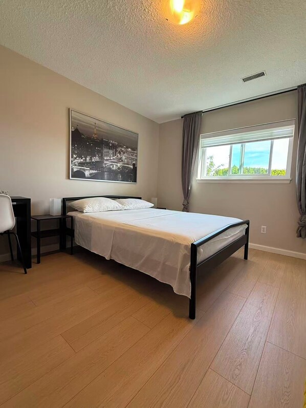 Un dormitorio con cama, escritorio, silla, ventana con cortinas e imagen colgada en la pared.