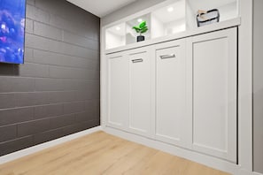 Une pièce avec un mur en briques grises, une armoire blanche à trois portes, une petite étagère avec une plante et un téléviseur fixé au mur.