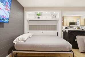 A modern hotel room with a bed, a TV mounted on the wall, a kitchenette, and a small plant on the shelf. - Cozy Downtown Townhouse with Patio (Kelowna)