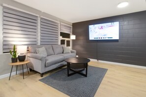 Ett modernt vardagsrum med en grå soffa, ett runt soffbord, en tv som är monterad på en mörk tegelvägg och ett litet sidobord med en lampa och en växt.