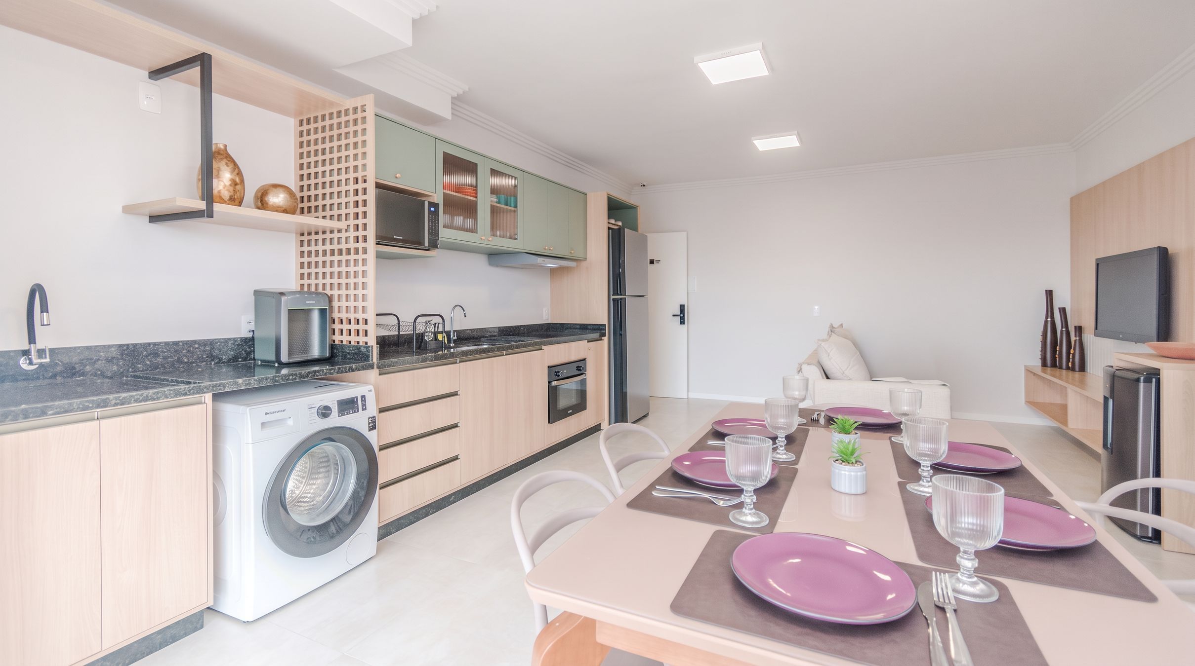 Een moderne keuken met een ingebouwde wasmachine, een eethoek met roze borden en glazen, en een aan de muur bevestigde televisie.