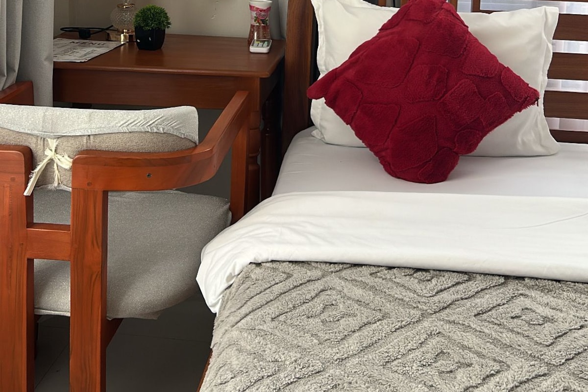 Quarto de hotel com cama, uma cadeira de madeira, mesa de cabeceira, luminária e janela com cortinas.