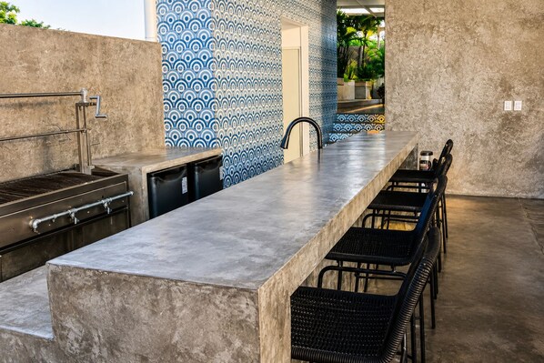 Una cocina exterior con una encimera larga, sillas negras y un revestimiento de pared estampado.