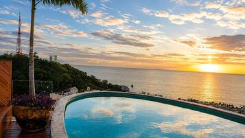 A curved infinity pool with a view of the sunset over the sea, surrounded by greenery and a wooden deck.