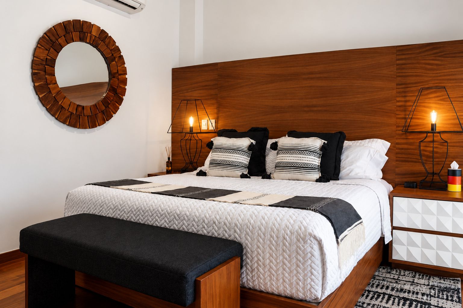 Un dormitorio moderno con cabecera de madera, un espejo redondo, lámparas de noche, una cama con ropa de cama estampada y una mesita de noche con una caja de pañuelos.