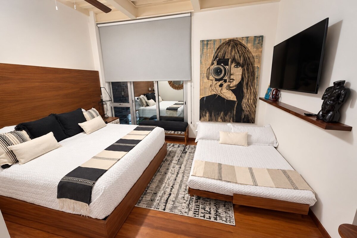 Un dormitorio moderno con dos camas, un ventanal grande, un ventilador de techo y un televisor montado.