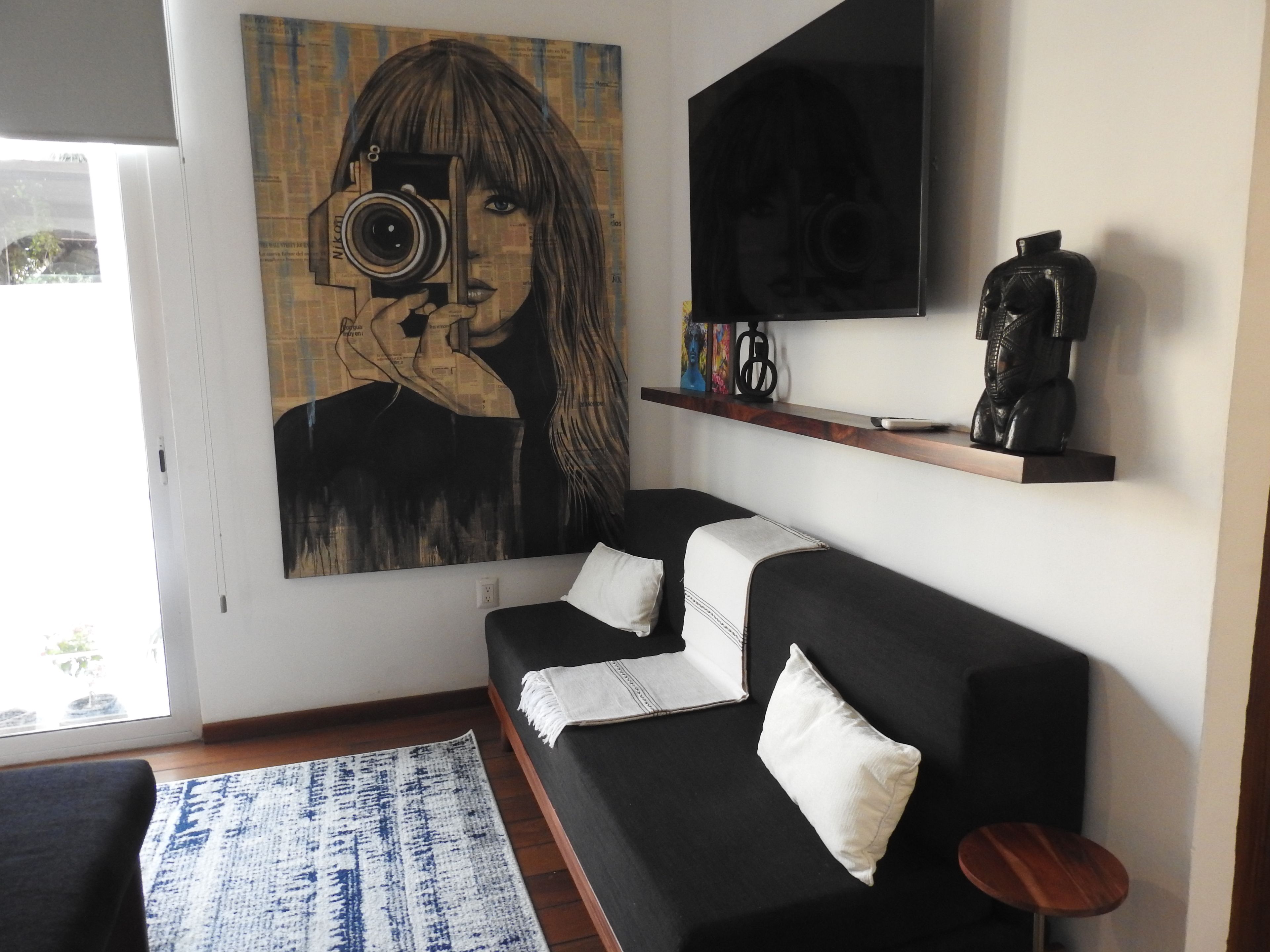 A living room with a black sofa, white cushions, a blue patterned rug, a coffee machine, and a large painting on the wall.
