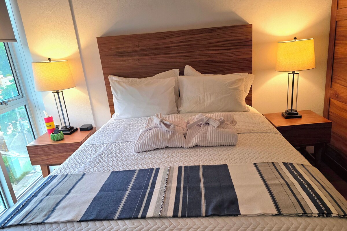A neatly made bed with white bedding, two pillows, and a pair of folded robes on a wooden headboard. There are two bedside lamps on wooden nightstands, and a window with a view of greenery outside.