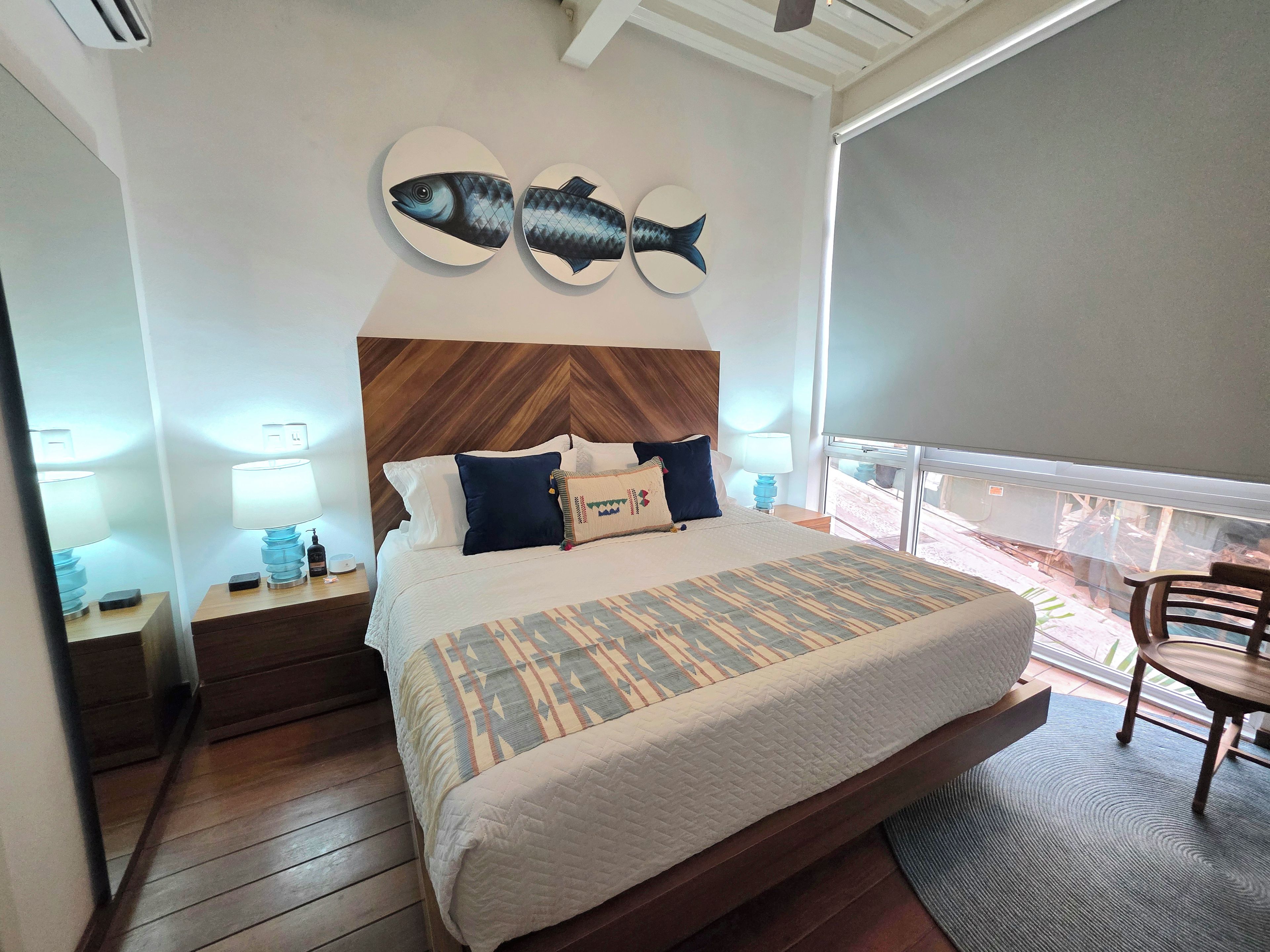 Habitación de hotel con cama, mesitas de noche, cabecero de madera, dos adornos de peces colgados en la pared, una ventana con vistas y un balcón con una silla.