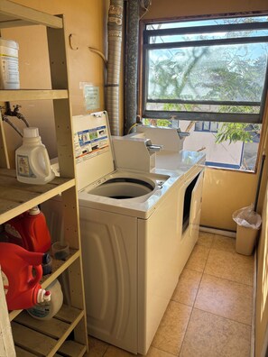A laundry room with a washing machine, shelves with cleaning supplies, a window with a view of trees, and a trash bin. - Historic 1-bedroom apartment in charming San Diego neighborhood with AC & Views (San Diego)