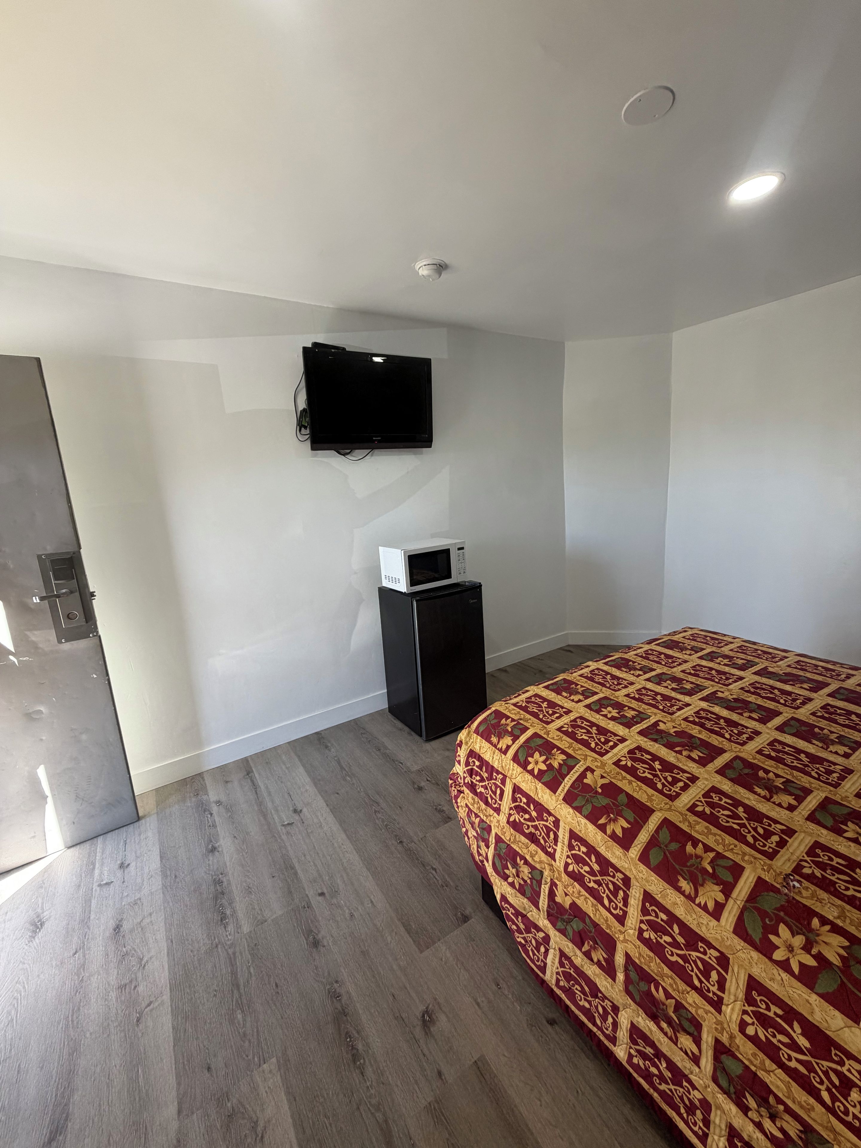 Un dormitorio con una cama, televisor montado en la pared, un refrigerador pequeño y piso de madera.