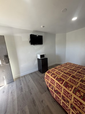 A bedroom with a bed, a television mounted on the wall, a small refrigerator, and a wooden floor. - deluxe motel (Pomona)