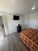 Un dormitorio con una cama, televisor montado en la pared, un refrigerador pequeño y piso de madera.