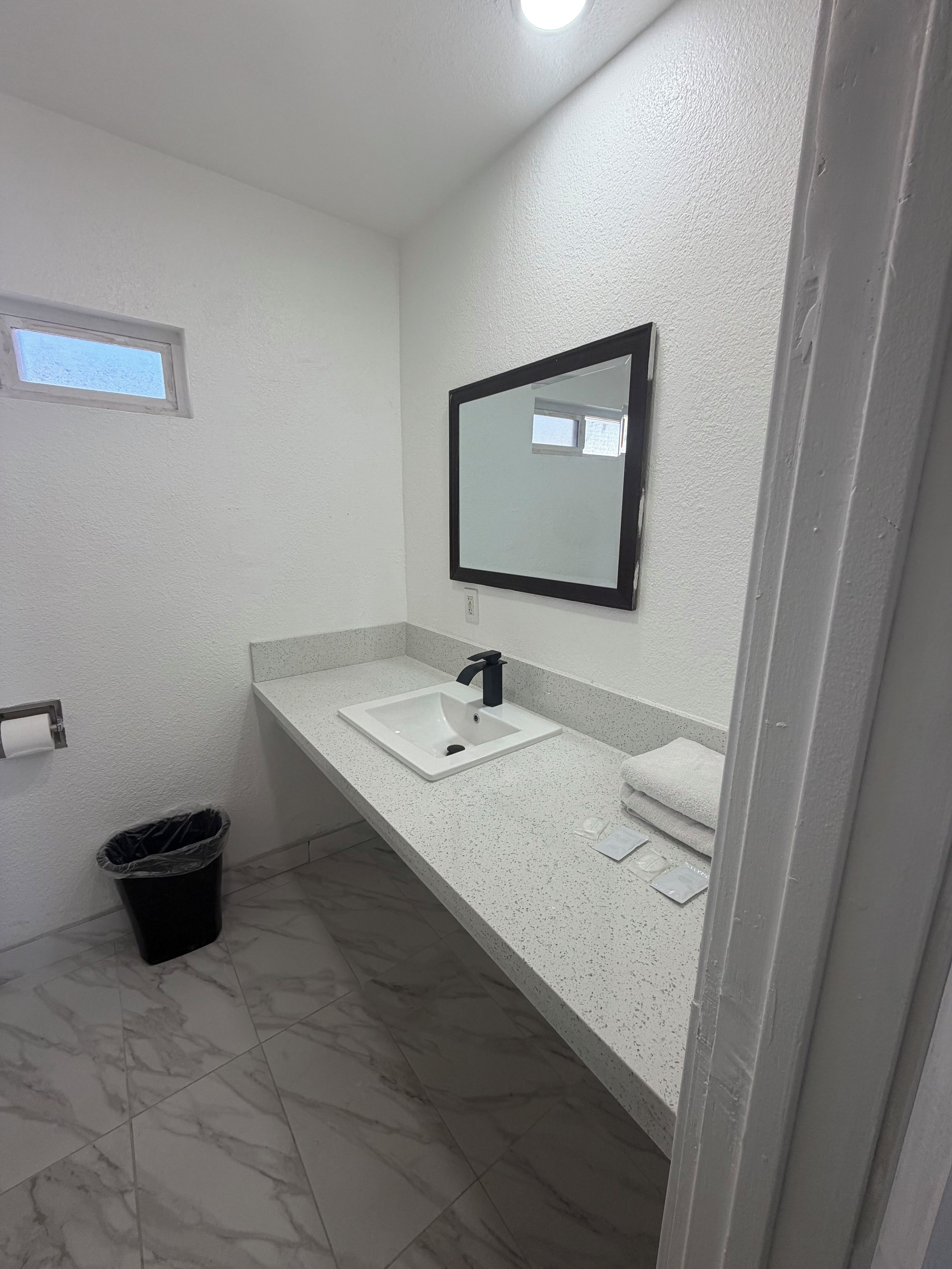Un baño con un lavamanos blanco, un espejo y un cubo de basura negro.
