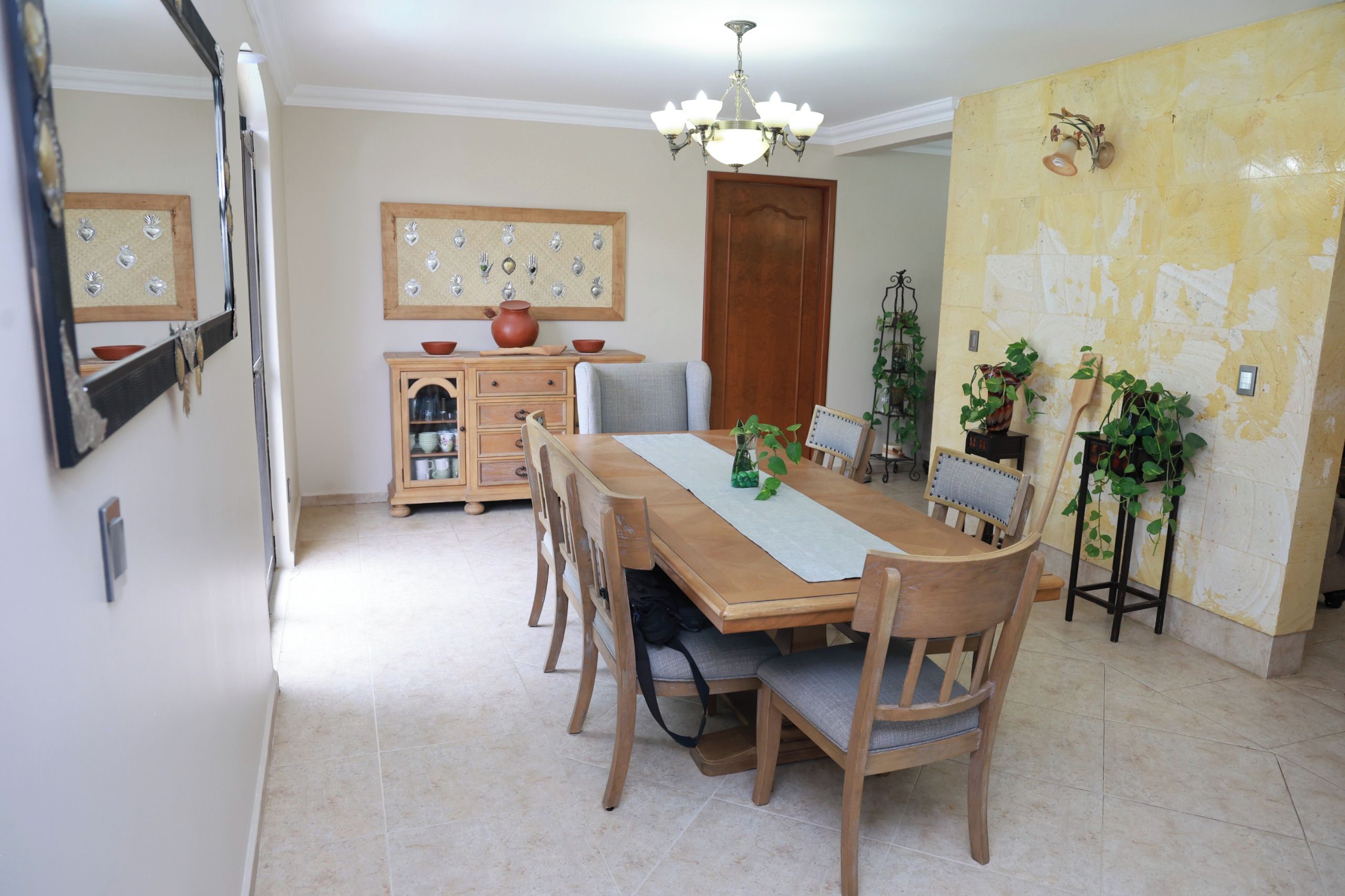 Un comedor con una mesa de madera, sillas, una araña, un armario con vajilla y una pared decorada.