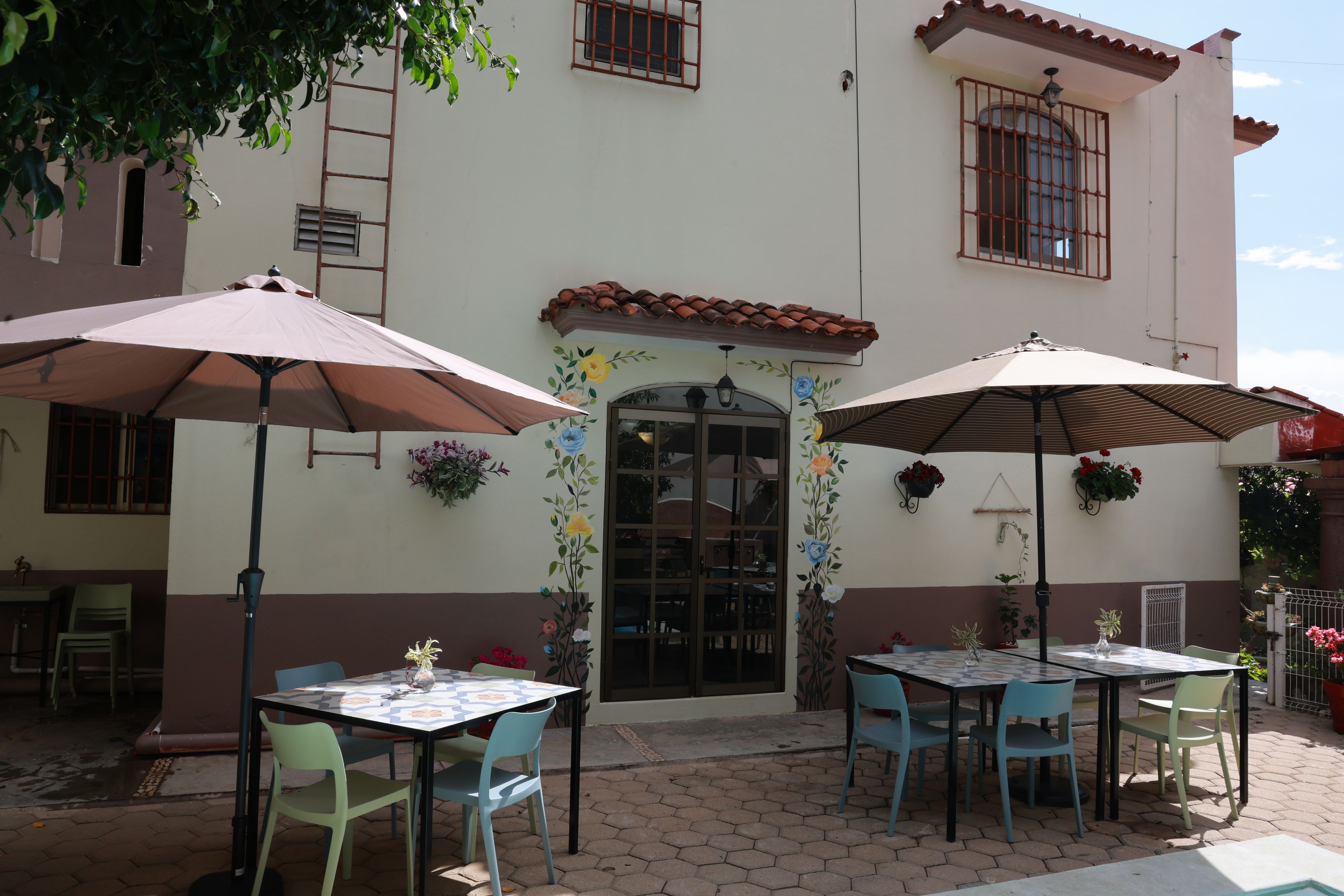Un patio al aire libre con mesas y sillas, una pared decorativa con motivos florales y un piso de baldosas.